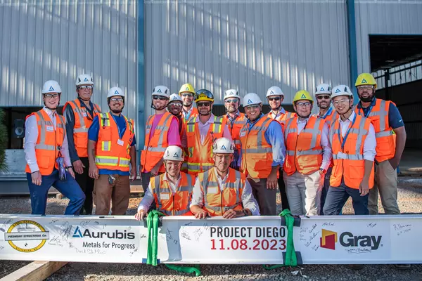 Foto: Aurubis: Eine Gruppe an Personen in Sicherheitswesten posieren vor einem grossen, bemalten Stahlträger für ein Gruppenfoto