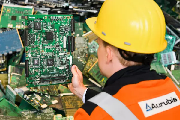 Foto: Aurubis: Mann mit gelbem Helm und orangener Aurubis Bekleidung ist von hinten zu sehen, er hält Microchip-Platten in der Hand