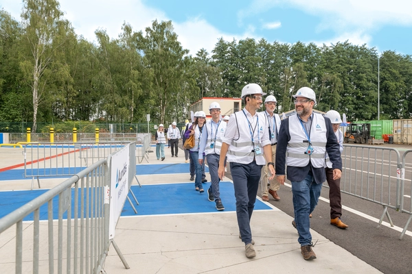 Foto: Aurubis: Mehrere Personen sind mit Helm und Weste bekleidet, sie gehen auf einer Strasse entlang zwischen Metallzaunen