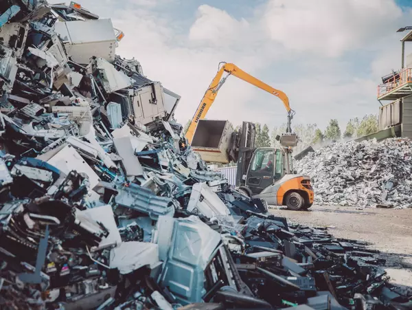 Foto: Aurubis: Ein gelber Bagger sortiert einen grossen Haufen Schrott auf einem Schrottplatz