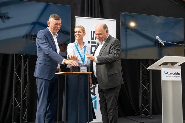 Foto: Aurubis: Zwei Maenner und eine Frau stehen auf einer Buehne, sie strecken die Hand aus