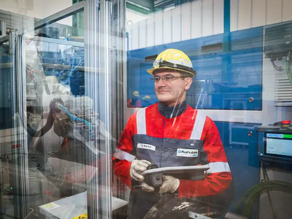 Foto: Aurubis: Ein Mann mit gelbem Helm und Aurubis Jacke steht vor einer Glaswand und hält ein Gerät in der Hand