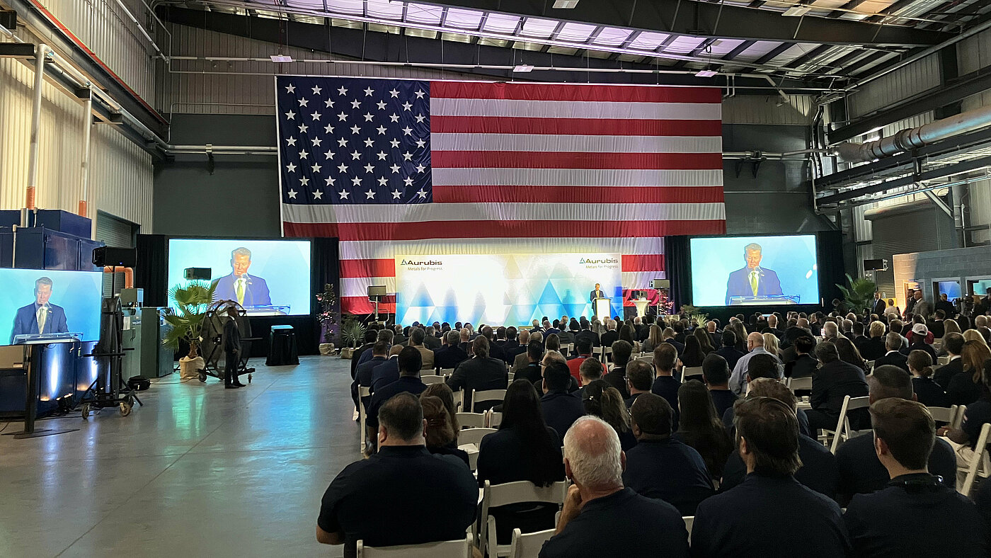 Foto: Aurubis: Mehrere Menschen sitzen auf Stühlen in einer Halle, vorne sind große Bildschirme und die USA Flagge